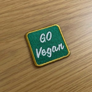 以下が含まれることがあります： 金色の縁取りのある緑色の刺繍パッチ。パッチには白い文字で「GO Vegan」と書かれています。