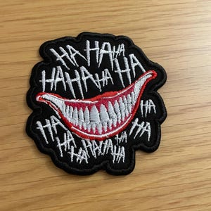 Pode incluir: Etiqueta bordada preta com uma boca sorridente vermelha e branca e o texto "HAHAHA" repetido ao redor da boca.