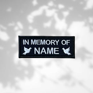 May include: A black rectangular patch with white embroidered text and two dove illustrations. The text reads "IN MEMORY OF" above "NAME". The patch is designed to be sewn onto clothing or fabric, a memorial item.