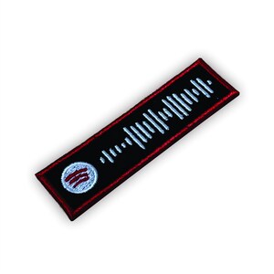 Op de afbeelding: Een zwarte en rode geborduurde patch met een wit geluidgolfontwerp en een witte cirkel met drie rode strepen in het midden. De patch is ongeveer 7,6 cm lang en 2,5 cm breed.