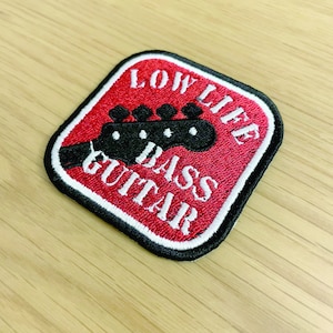 Peut inclure: Patch brodé carré rouge avec du texte blanc qui dit "Low Life Bass Guitar" et une illustration noire de tête de basse.