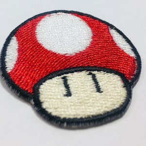 Könnte beinhalten: Ein gestickter Pilz-Patch in Rot und Weiß mit schwarzem Umriss. Der Pilz hat einen weißen Stiel mit zwei schwarzen Punkten.