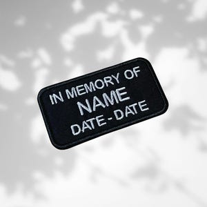 May include: Black rectangular patch embroidered with white thread. The text "IN MEMORY OF NAME DATE-DATE" is centered on the patch. This patch is likely a memorial item, possibly for clothing or accessories.