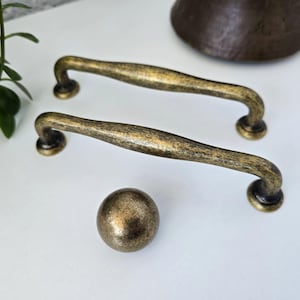 May include: Antique brass cabinet hardware set. The set includes two curved handles and a round knob. The hardware has a distressed finish with dark accents, suggesting an aged appearance. The handles are approximately 15 cm long.