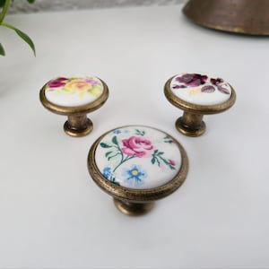 Op de afbeelding: Drie decoratieve kastknoppen met bloemmotieven. De knoppen hebben een wit keramisch oppervlak met kleurrijke bloempatronen, waaronder roze rozen en blauwe accenten. De knoppen hebben een bronzen metalen basis.