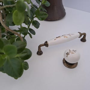 Könnte beinhalten: Ein weißer Keramik-Möbelgriff und ein passender runder Möbelknopf, beide mit einem zarten goldenen und grauen Blumenmuster verziert. Die Beschläge verfügen über Sockel und Befestigungspunkte aus antikem bronzefarbenem Metall als Kontrast.