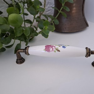 Puede incluir: Un tirador de armario de cerámica blanca con un diseño floral en rosa y azul. El tirador tiene extremos metálicos de color bronce. El tirador está diseñado para muebles y tiene un estilo clásico y decorativo.