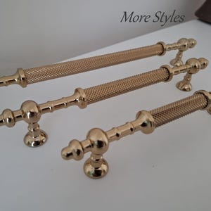 May include: Three gold-colored metal cabinet pulls arranged in a row. Each pull features a central textured, knurled cylindrical grip section flanked by smooth, rounded decorative ends and two mounting posts. The text More Styles is visible.