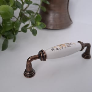 Könnte beinhalten: Ein Schrankgriff mit einem weißen Keramikmittelteil mit goldenem Blumendesign. Die Enden und gebogenen Stützen sind dunkelbronze. Der Griff ist etwa 13 cm lang.