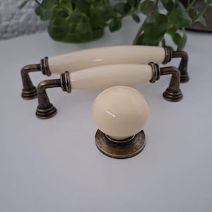 Può includere: Maniglie per mobili in ceramica color crema e un pomello abbinato con ferramenta in bronzo antico. Le maniglie sono ovali e il pomello è sferico. La ferramenta ha un aspetto vintage, invecchiato, che suggerisce uno stile classico o retrò.