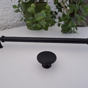 Può includere: Un set di maniglia e pomello per armadi neri abbinati. La maniglia è una barra lunga e dritta con estremità decorative. Il pomello è rotondo e piatto. Entrambi i pezzi sono in metallo nero opaco. Ferramenta per armadi e cassetti.
