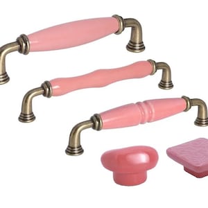 Op de afbeelding: Een verzameling roze keramische kastbeslagen met antiek messingkleurige metalen accenten. De set bevat drie verschillende handgreepstijlen en twee knopstijlen. De handgrepen hebben een gebogen ontwerp, terwijl de knoppen rond en vierkant zijn.