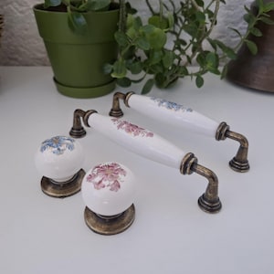 Puede incluir: Juego de dos tiradores de armario de cerámica blanca y dos pomos redondos a juego. Los tiradores y pomos presentan diseños florales en rosa y azul, con detalles metálicos color latón antiguo. Para armarios de cocina o baño.