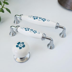 Puede incluir: Tiradores y pomo de armario de cerámica blanca con diseño floral azul. Los tiradores tienen forma curva con herrajes metálicos cromados. El pomo es redondo con un patrón floral a juego. Herrajes decorativos para muebles.