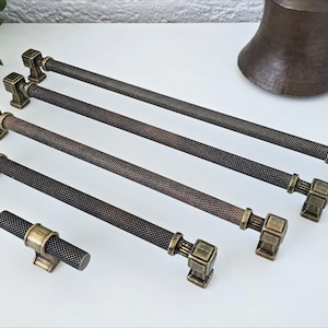 Peut inclure: Ensemble de poignées de meubles de couleur bronze et un bouton. Les poignées ont une barre cylindrique texturée avec des embouts carrés. Le matériel est disposé sur une surface blanche, mettant en valeur le design et la finition décoratifs.