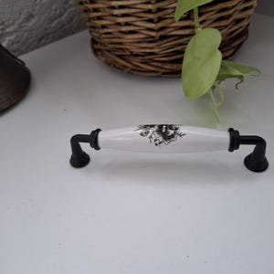 May include: A cabinet pull featuring a white ceramic center handle with a black floral pattern. The handle is flanked by two matte black metal mounting brackets on each side, which connect to the ends of the ceramic piece with circular bases.