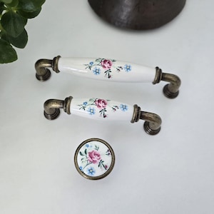 May include: Two white ceramic drawer pulls and one matching round knob, each featuring a floral pattern with pink roses and blue flowers. The hardware has an antique bronze-toned metal finish on the ends and circular mounting bases.
