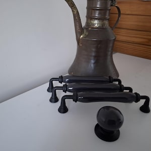 Op de afbeelding: Drie zwarte keramische kastgrepen met matzwart metalen beslag en een bijpassende ronde zwarte keramische kastknop. De grepen hebben een glad, langwerpig cilindrisch middenstuk met getrapte ronde voetjes aan elk uiteinde van de handgreep.