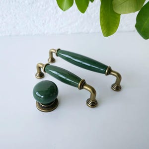Peut inclure: Un ensemble de trois pièces de quincaillerie d'armoire en céramique vert foncé. L'ensemble comprend deux poignées allongées et un bouton rond, le tout avec des accents métalliques de couleur laiton. La quincaillerie est conçue pour les meubles et les portes d'armoires.