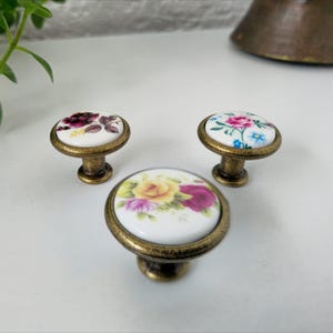 Op de afbeelding: Drie decoratieve kastknoppen met bloemmotieven. De knoppen hebben een wit keramisch oppervlak met kleurrijke rozenpatronen in roze, gele en paarse tinten. Ze zijn gemonteerd op antiek messingkleurige bases. De knoppen zijn ontworpen voor meubels.