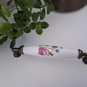 Pode incluir: Puxador de arm&aacute;rio de cer&acirc;mica branca com um design floral com rosas cor-de-rosa e flores azuis. O puxador tem ferragens de metal com cor de bronze antigo em cada extremidade. Um detalhe decorativo para m&oacute;veis.