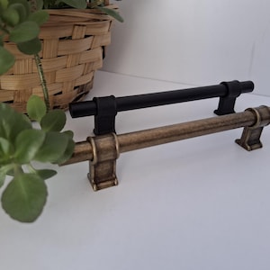 May include: Two cabinet pulls with a cylindrical bar design and square mounting posts. One pull features a matte black finish, while the other is finished in an antique brass tone. Both hardware pieces have a horizontal orientation.