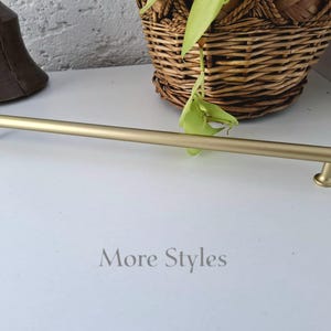 May include: A long, cylindrical cabinet pull handle with a brushed gold finish. The hardware features a smooth, straight bar design supported by two small, circular pedestals at each end. The text "More Styles" is printed in gray below the handle.