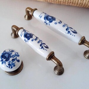 May include: A set of cabinet hardware featuring a white porcelain knob and two handles. Each piece is adorned with a blue floral design and accented with antique brass-colored metal. The handles measure approximately 6 inches in length.