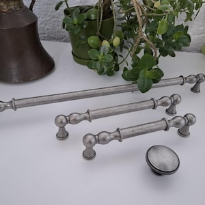 May include: A collection of cabinet hardware featuring three elongated metal drawer pulls and one round knob. Each piece has a brushed, weathered silver finish with a textured surface, rounded ends, and a central cylindrical bar design.