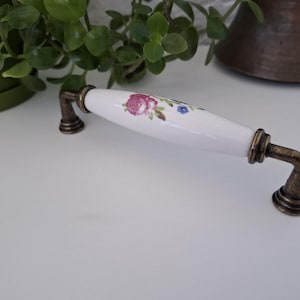 Pode incluir: Um puxador de arm&aacute;rio de cer&acirc;mica branca com um design floral em rosa, roxo e azul. A al&ccedil;a tem ferragens de metal cor de bronze. O puxador &eacute; um elemento decorativo para m&oacute;veis.