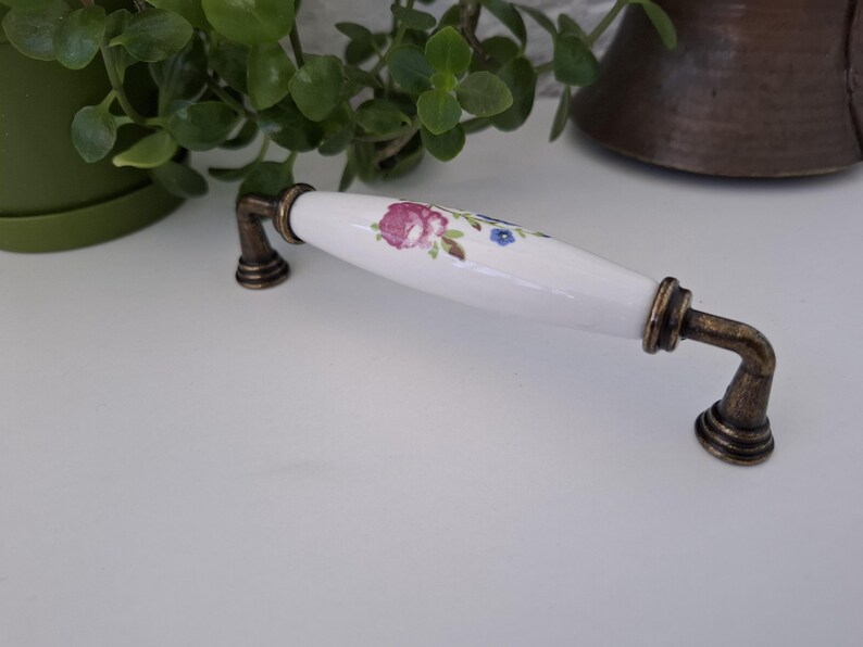 Pode incluir: Um puxador de arm&aacute;rio de cer&acirc;mica branca com um design floral em rosa, roxo e azul. A al&ccedil;a tem ferragens de metal cor de bronze. O puxador &eacute; um elemento decorativo para m&oacute;veis.