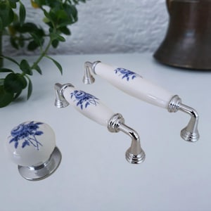 May include: Two cabinet pulls and one matching round cabinet knob, all featuring a white ceramic surface with a blue floral pattern and polished silver-tone metal hardware. The hardware includes ribbed circular bases for secure mounting.
