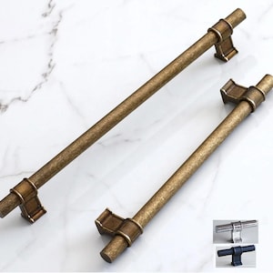 May include: Two antique bronze cabinet pulls. The pulls are cylindrical with decorative end pieces. The pulls are shown on a white marble surface. Additional pulls in silver and black are shown in the lower right corner.