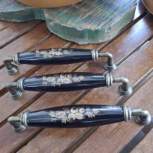 Puede incluir: Tres tiradores de cajón con acabado negro brillante y un patrón floral central de color crema. Cada tirador presenta una forma alargada y redondeada con herrajes de metal en tono latón antiguo en ambos extremos para su montaje.