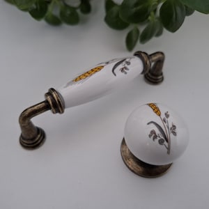 May include: A white ceramic cabinet handle and a matching round cabinet knob, both featuring a painted yellow wheat stalk and grey foliage design. The hardware includes antique bronze-toned metal bases and curved mounting brackets for attachment.