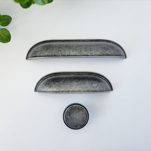May include: Three pieces of antique-style cabinet hardware. Two are half-moon shaped pulls, and one is a round knob. The hardware has a distressed, mottled grey finish, suggesting an aged appearance. The items are arranged on a white surface.