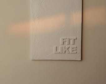 May include: A rectangular white canvas with textured surface, mounted on a wall. The words "FIT LIKE" are embossed in the lower right corner. The wall is a light beige color, with a warm light shining on the canvas.