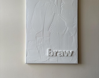 May include: White textured canvas art with the word "braw" in raised lettering. The artwork is rectangular and mounted on a wall. The canvas has a thick, layered texture.