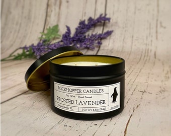 May include: A black soy wax candle in a tin with a gold rim and lid. The label reads "Rockhopper Candles Frosted Lavender" with a penguin logo. The candle weighs 6.5oz (184g). A sprig of purple lavender is in the background.