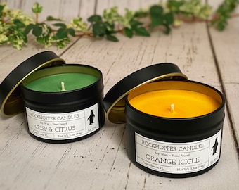 May include: Two soy wax candles in black tins with gold lids. The candles are labeled "Rockhopper Candles" and have a penguin logo. The candle on the left is green and labeled "Crisp & Citrus". The candle on the right is yellow and labeled "Orange Icicle".