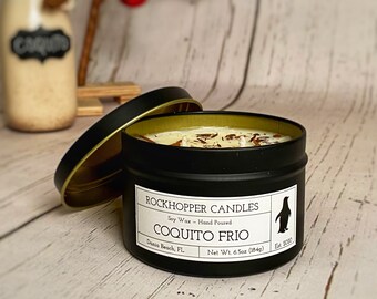 May include: A black metal candle tin with a gold rim and lid, labeled "COQUITO FRIO" by Rockhopper Candles. The candle contains a creamy wax with brown accents and weighs 6.5 oz (184g). A penguin logo is on the label.