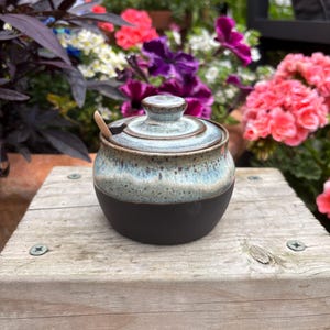 May include: A small ceramic sugar bowl with a lid and a wooden spoon. The bowl features a matte black base and a speckled, light blue and cream glazed upper section. The lid has a matching blue and cream glaze with a central knob handle.