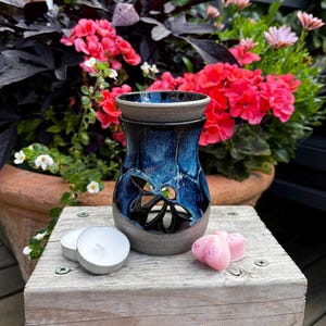 May include: A ceramic wax warmer featuring a deep blue and black reactive glaze with a cutout floral pattern. The base is a matte grey, and it is accompanied by two white tea light candles and two small pink heart-shaped wax melts.