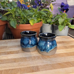 May include: Two handmade ceramic mugs with a blue glaze over a tan base. The mugs have a unique, textured appearance and a dark blue interior. The mugs are displayed on a wooden chopping board, with potted flowers in the background.