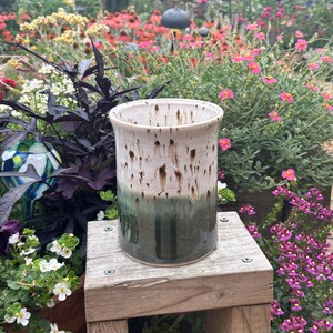 May include: A cylindrical ceramic utensil holder featuring a two-tone glaze. The top half is an off-white colour with vertical brown drip patterns, while the bottom half is a solid, deep forest green. The rim is smooth and rounded.