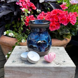 May include: A ceramic wax warmer with a glossy blue and black reactive glaze finish, featuring cut-out shapes around the base. Two round white tea light candles and two small pink heart-shaped wax melts are positioned in front of the warmer.