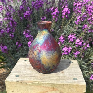 May include: A handmade ceramic vase with an iridescent finish. The vase has a rounded base and a slender neck. It displays a mix of colours, including red, yellow, blue, and purple. It is placed on a wooden surface, with purple flowers in the background.