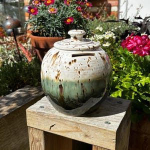 May include: A round ceramic coin bank with a removable lid and a horizontal slot. The upper portion is a cream colour with brown speckles, while the lower portion features a dark green glaze that drips downwards, creating a smooth, glossy finish.