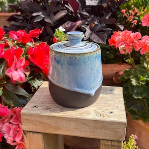 May include: A ceramic jar with a lid featuring a two-tone design. The upper portion and lid are finished in a mottled light blue glaze with darker blue speckles, while the lower portion is a solid, matte dark charcoal grey ceramic base.