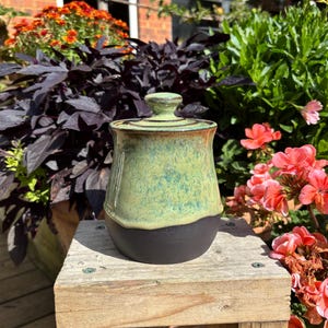 May include: A ceramic jar with a lid featuring a two-tone glaze. The upper portion has a mottled light green and teal finish, while the lower base is a solid matte black. The jar has a rounded body and a small knob handle on the lid.